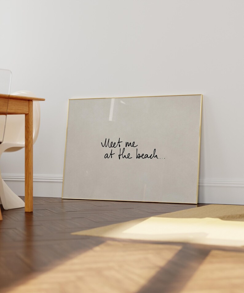 Puede incluir: Un cuadro enmarcado con el texto "Meet me at the beach..." en estilo manuscrito. El marco es dorado y el fondo es de color beige claro. El cuadro est&aacute; apoyado contra una pared blanca.