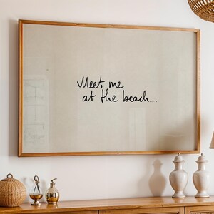 Puede incluir: Una impresi&oacute;n enmarcada con el texto escrito a mano "Meet me at the beach" en escritura negra. El marco es de madera marr&oacute;n claro. La impresi&oacute;n cuelga sobre un mueble de madera con objetos decorativos.