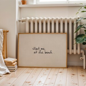 Puede incluir: Una impresi&oacute;n art&iacute;stica rectangular enmarcada con el texto "Meet me at the beach..." en escritura negra. La impresi&oacute;n tiene un fondo beige claro y un marco de madera natural. La impresi&oacute;n se exhibe en una habitaci&oacute;n con un radiador, una planta en maceta y una pila de libros.