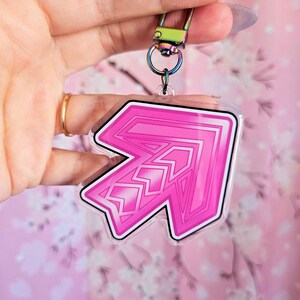 Puede incluir: Un llavero en forma de flecha rosa y blanca con un contorno negro. El llavero tiene un cierre plateado y un respaldo de plástico transparente. El diseño de la flecha presenta una serie de zigzags. El fondo es un estampado floral rosa.