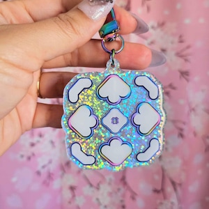 May include: A square-shaped keychain with a holographic glitter background and white cloud-like shapes. The keychain has a rainbow-colored clasp and a silver ring. The design includes a small square in the center with a house icon.