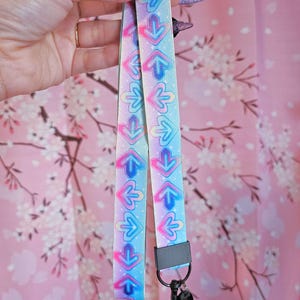 May include: A pastel-colored lanyard with a repeating arrow pattern in pink, blue, and purple. The lanyard has a black clasp and key ring. The background is a pink floral pattern.