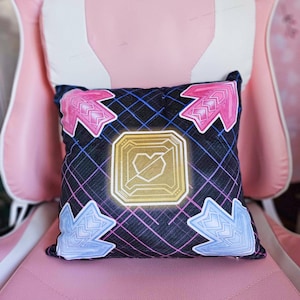 May include: A decorative pillow with a black background and a grid pattern. The pillow features four arrow-shaped designs in pink and blue, surrounding a gold square with a heart symbol. The pillow is placed on a pink chair.