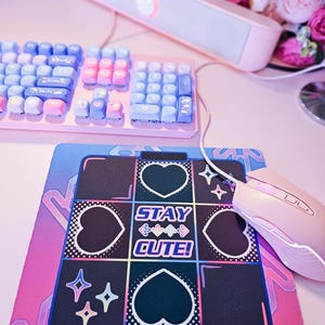 May include: A pink and blue gaming mouse pad with a black and white design. The design features hearts, stars, and the text "STAY CUTE!" in a neon pink font.