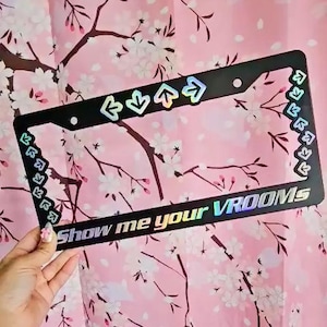 Puede incluir: Un marco de matrícula negro con flechas iridiscentes y el texto "Show me your VROOMs". El marco está sobre un fondo rosa con un patrón floral blanco.