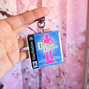 May include: A blue and pink iridescent keychain featuring a video game cover for "Dance Dance Revolution" for the PlayStation. The cover features a pink silhouette of a person dancing on a blue platform.