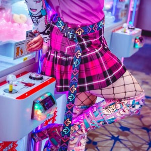Peut inclure: Une personne portant une jupe à carreaux rose et noire avec une ceinture noire ornée de cœurs et de flèches multicolores. La personne porte également des collants en résille noirs et des bottes cuissardes avec un imprimé coloré d'inspiration anime.