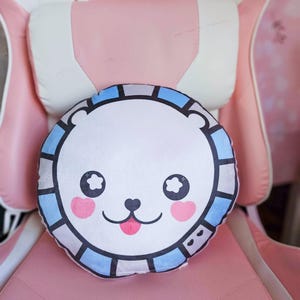 Puede incluir: Una almohada redonda y blanca con un diseño de cara de oso de dibujos animados. El oso tiene ojos negros, mejillas rosas y una lengua rosa. La almohada está bordeada con cuadrados azules y grises, sobre una silla rosa.