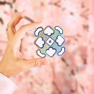 Puede incluir: Una pegatina holográfica con un patrón repetitivo de nubes blancas y líneas curvas. La pegatina tiene un acabado brillante.