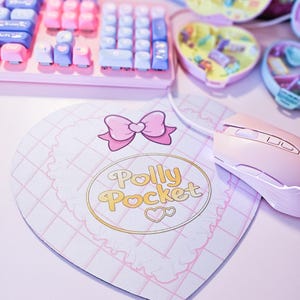 May include: A heart-shaped mousepad with a pink bow and the words "Polly Pocket" in gold lettering. A pink computer mouse and keyboard are also visible, along with miniature Polly Pocket toys.