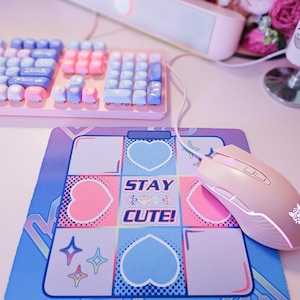 May include: A pink and blue mouse pad with a white border and a graphic design of hearts, stars, and the text "STAY CUTE!" A pink gaming mouse is on the mouse pad.