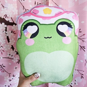 May include: Green frog plush toy with big eyes and a pink and white flower hat. The frog has a happy expression and rainbow-colored pupils.