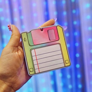 May include: A colorful, retro-style floppy disk-shaped notepad. The notepad features a pink, green, and yellow color scheme with a lined paper section and a red vertical line. It has a small hole at the top for hanging.