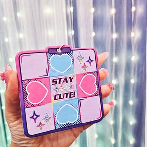 May include: A pink and blue square card with a white border and a white heart in each corner. The card has a pink heart in the centre with the text "STAY CUTE!" in white letters. The card has a white border and a white heart in each corner.