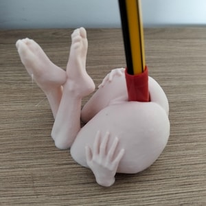May include: A light pink figurine of a person in a reclining pose, with legs and a hand visible. A yellow and black pencil is inserted into the figurine. The figurine is on a wooden surface.