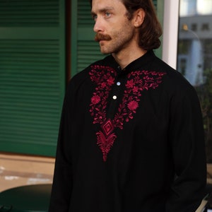 Puede incluir: Camisa negra de manga larga con cuello mandarín y bordado floral decorativo en tonos rojos. El bordado está centrado en el pecho y alrededor del cuello. La camisa tiene tres botones blancos.
