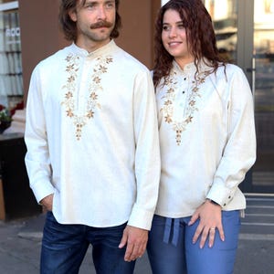 Puede incluir: Dos personas con camisas crema de manga larga a juego con intrincados bordados florales marrones alrededor del cuello. Una persona lleva vaqueros azul oscuro, la otra pantalones azul claro. Cuello mandar&iacute;n.