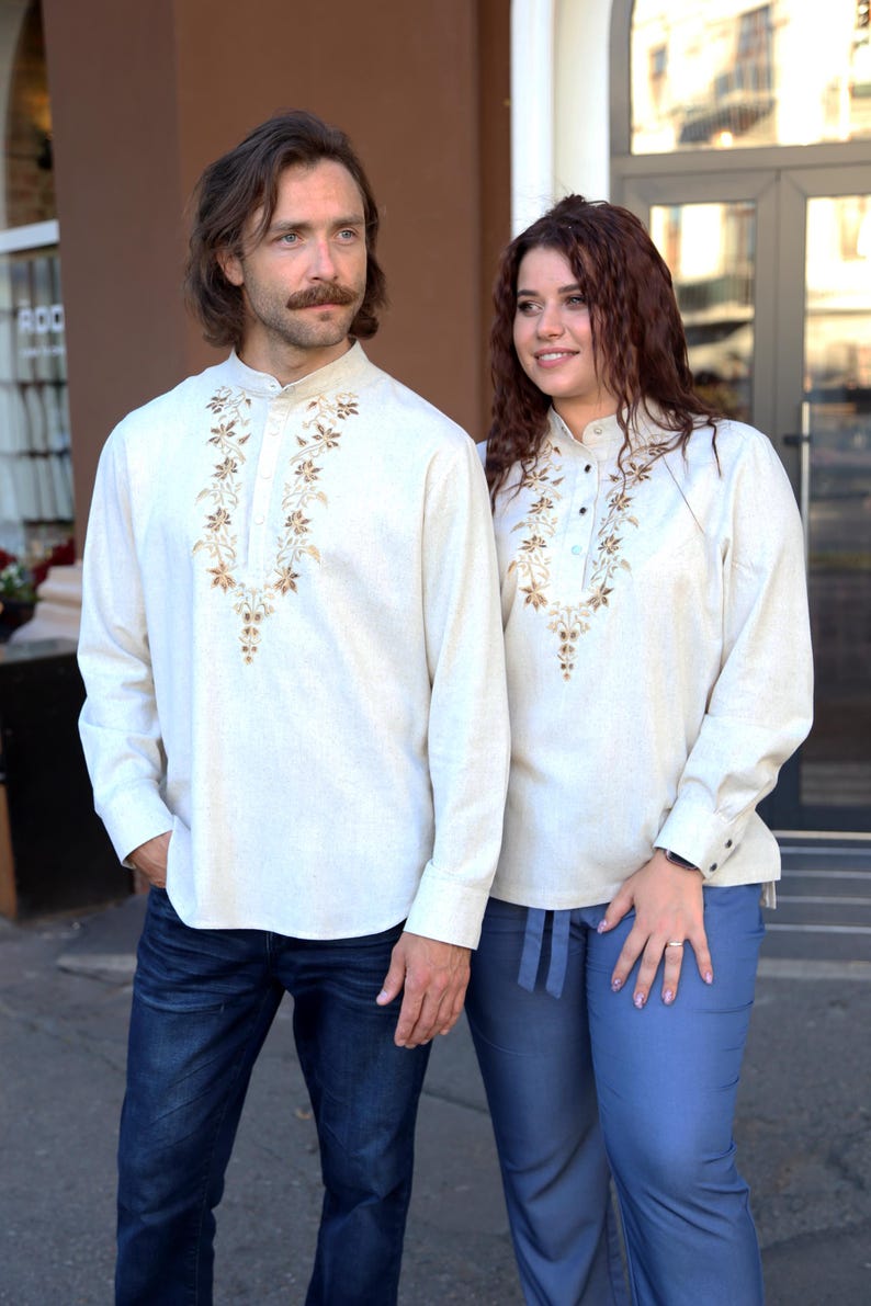 Puede incluir: Dos personas con camisas crema de manga larga a juego con intrincados bordados florales marrones alrededor del cuello. Una persona lleva vaqueros azul oscuro, la otra pantalones azul claro. Cuello mandar&iacute;n.