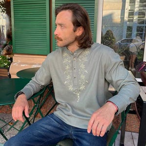 Puede incluir: Una camisa verde claro de manga larga con bordados florales alrededor del cuello y en la parte delantera. La camisa se combina con vaqueros azules. La persona est&aacute; sentada en una silla verde.