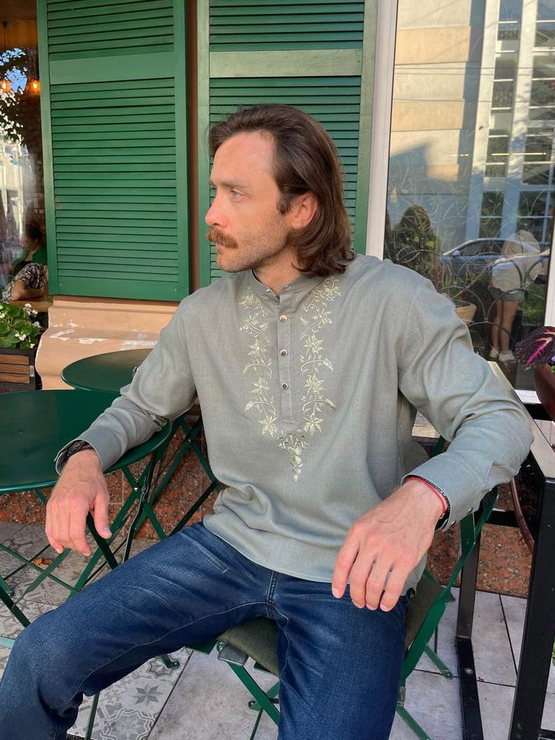 Puede incluir: Una camisa verde claro de manga larga con bordados florales alrededor del cuello y en la parte delantera. La camisa se combina con vaqueros azules. La persona est&aacute; sentada en una silla verde.