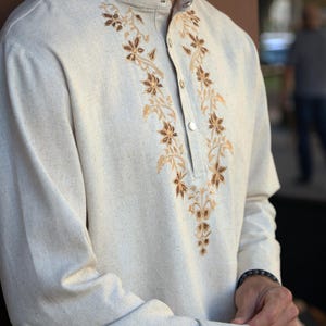 Puede incluir: Camisa beige de manga larga con cuello mandar&iacute;n y abotonadura frontal. Presenta un intrincado bordado floral en tonos marr&oacute;n y dorado a lo largo del escote. La tela tiene una apariencia texturizada.