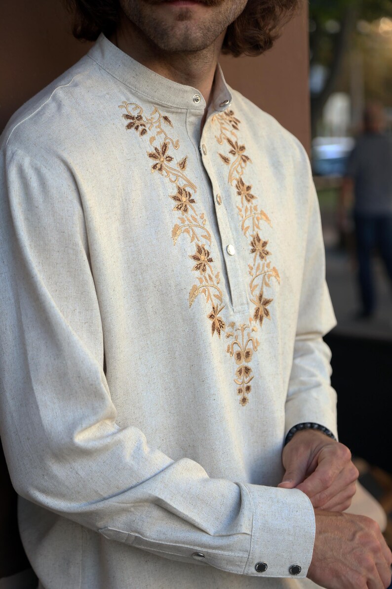 Puede incluir: Camisa beige de manga larga con cuello mandar&iacute;n y abotonadura frontal. Presenta un intrincado bordado floral en tonos marr&oacute;n y dorado a lo largo del escote. La tela tiene una apariencia texturizada.