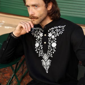 May include: A black long-sleeved shirt with a white embroidered floral design on the front. The shirt has a mandarin collar and a single button closure.