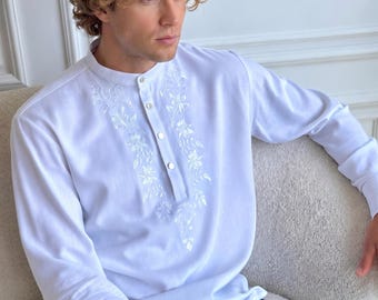 May include: A white long-sleeved shirt with a mandarin collar and a button-down placket. The shirt features white floral embroidery along the front placket and cuffs with metallic buttons. The fabric appears smooth and lightweight in construction.