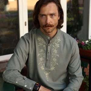 Puede incluir: Una camisa verde claro con bordados florales alrededor del cuello y en la parte delantera. La camisa tiene cuello mandar&iacute;n y pu&ntilde;os abotonados. La persona lleva un reloj negro y una pulsera.
