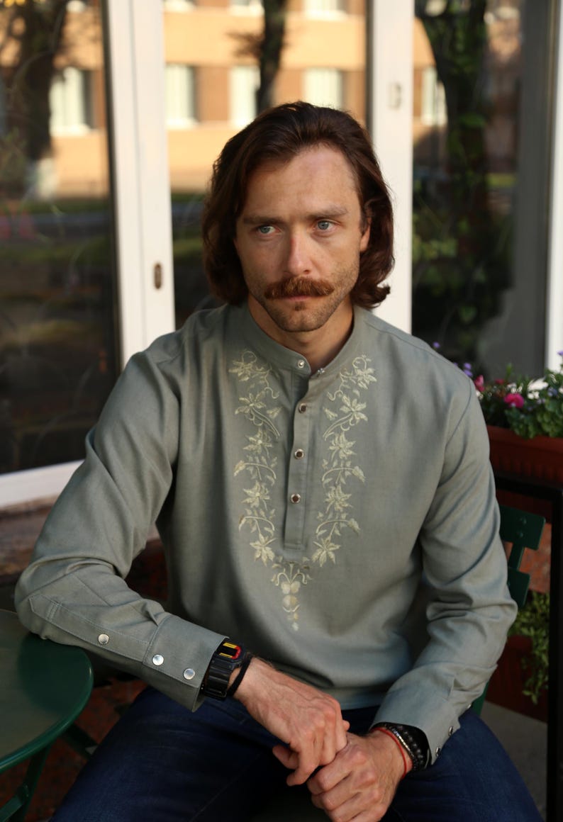 Puede incluir: Una camisa verde claro con bordados florales alrededor del cuello y en la parte delantera. La camisa tiene cuello mandar&iacute;n y pu&ntilde;os abotonados. La persona lleva un reloj negro y una pulsera.