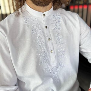 Puede incluir: Camisa blanca de manga larga con cuello mao y tapeta abotonada. La camisa presenta delicados bordados florales en la parte delantera. Hecha de una tela ligera.