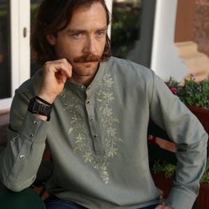 Puede incluir: Una camisa verde salvia con mangas largas y cuello mandar&iacute;n. La camisa presenta bordados florales intrincados en la parte delantera y en los pu&ntilde;os. La camisa est&aacute; abotonada y tiene un corte relajado, adecuada para uso casual.