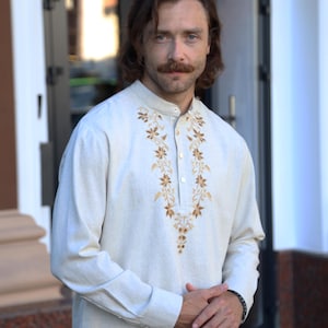 May include: A long-sleeved, off-white linen shirt with a mandarin collar and intricate gold floral embroidery down the front. The shirt has a button-up placket and is worn by a person.