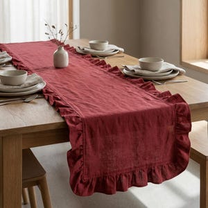 May include: A burgundy linen table runner with ruffled edges. The runner is placed on a wooden table, set with neutral-toned plates, bowls, and silverware. A small vase with dried stems sits in the center.