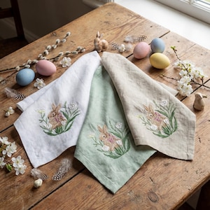 Può includere: Tre asciugamani pasquali in lino bianco, verde e beige, ciascuno ricamato con un coniglietto in un ambiente floreale. La scena comprende uova dipinte, coniglietti decorativi e ramoscelli di fiori su un tavolo di legno rustico.