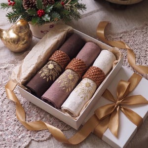 Puede incluir: Una caja de regalo blanca que contiene tres servilletas de tela enrolladas en color marrón oscuro, malva y crema. Cada servilleta está sujeta con un servilletero de ratán trenzado y presenta un diseño floral dorado bordado.