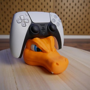 Könnte beinhalten: Ein weißer und schwarzer PlayStation 5-Controller mit orangefarbenen und blauen Details sitzt auf einem orangefarbenen, dinosaurierförmigen Ständer.