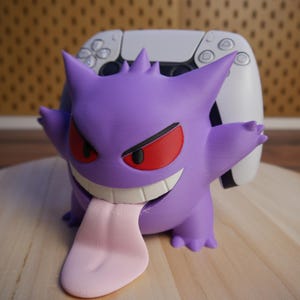 Könnte beinhalten: Eine lila Gengar-Figur mit roten Augen und einer rosa Zunge, die vor einem weißen Gamecontroller platziert ist. Die Figur hat ein bedrohliches Grinsen und steht auf einer Holzoberfläche.
