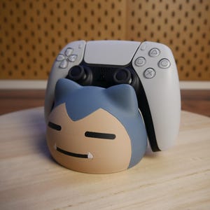 Könnte beinhalten: Ein weißer PlayStation 5-Controller sitzt auf einem blauen und braunen Pokemon-Charakter-Ständer. Der Ständer ist in Form eines Pokemon-Charakters mit einem blauen Hut und einem braunen Gesicht gestaltet.