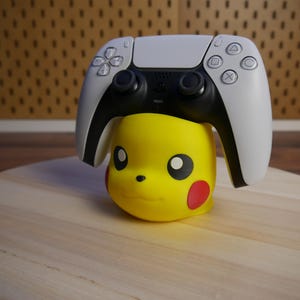 Könnte beinhalten: Ein weißer und schwarzer Videospiel-Controller sitzt auf einer gelben Pikachu-Figur. Der Controller hat eine weiße Vorderseite mit schwarzen Knöpfen und eine schwarze Rückseite.