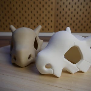 Könnte beinhalten: Zwei 3D-gedruckte Pokémon-Schädelmodelle, eines ist weiß und das andere ist hellbraun. Die Modelle stehen auf einer Holzoberfläche.