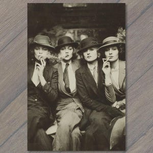 May include: A black and white vintage photograph depicting four individuals wearing tailored suits, pinstriped trousers, and felt fedora hats. Each person is dressed in formal attire with collared shirts and ties, while holding cigarettes.