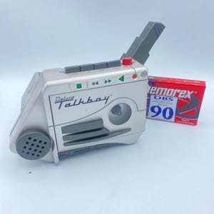 May include: A silver Deluxe Talkboy tape recorder with a gray handle and red button. The Talkboy has a speaker, play, rewind, and fast forward buttons. A red and white cassette tape is next to the Talkboy.