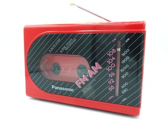 Puede incluir: Una grabadora de casetes de radio FM/AM Panasonic roja. La radio tiene un panel negro con texto blanco y una ventana transparente que muestra la pletina de casetes. La antena está extendida. La radio tiene las palabras "FM AM" impresas.