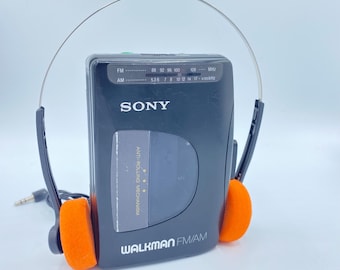 Puede incluir: Un reproductor de casetes Sony Walkman negro con una diadema plateada y almohadillas para los oídos de color naranja. El reproductor tiene una pantalla digital con el texto "FM" y "AM" y los rangos de frecuencia para cada banda. El texto "ANTI-ROLLING MECHANISM" está impreso en el reproductor. El texto "WALKMAN FM/AM" está impreso en la parte inferior del reproductor.