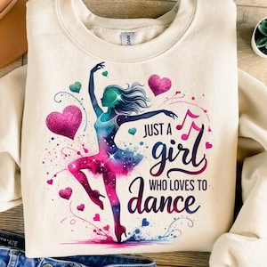Può includere: Felpa color crema con un disegno ad acquerello di una ballerina nei toni del blu, rosa e viola. È incluso anche il testo "Just a girl who loves to dance". Il design incorpora cuori e note musicali.
