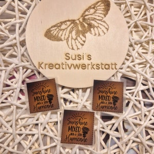 May include: Three square iron-on patches with a brown background and white text that reads "Sunshine Mixed with a little Hurricane". The patches are on a white wicker surface with a wooden circle behind them that has the text "Susi's Kreativwerkstatt" burned into it.