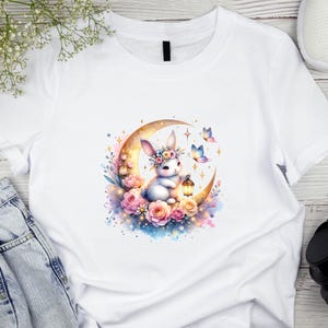 May include: White t-shirt with a watercolour illustration of a bunny wearing a floral crown, seated on a crescent moon. The design includes pink roses, butterflies, and a lantern, with a soft, pastel colour palette.