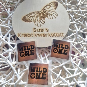 Puede incluir: Tres parches termoadhesivos marrones y negros con el texto "WILD ONE" en una fuente retro. Cada parche presenta un pequeño diseño floral.