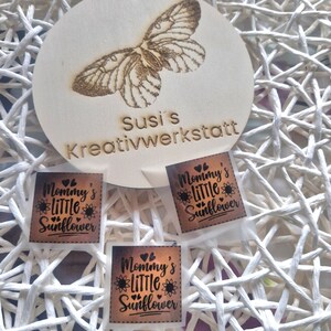 Könnte beinhalten: Ein Holzschild mit einer Schmetterlingsgravur und dem Text "Susi's Kreativwerkstatt". Darunter befinden sich drei kleine, quadratische Etiketten mit dem Text "Mommy's Little Sunflower" und einer Sonnenblumen-Grafik.